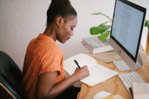 Woman in an orange shirt studying at a desk with a computer. She is practicing our three tips for effective studying.